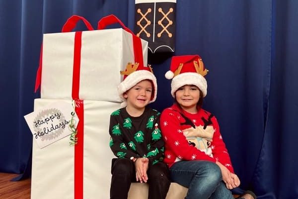 Two children in festive sweaters and Santa hats sit on a small white box in front of a large wrapped gift. The gift is decorated with red ribbon and a Happy Holidays tag. A blue curtain and holiday decoration are in the background.