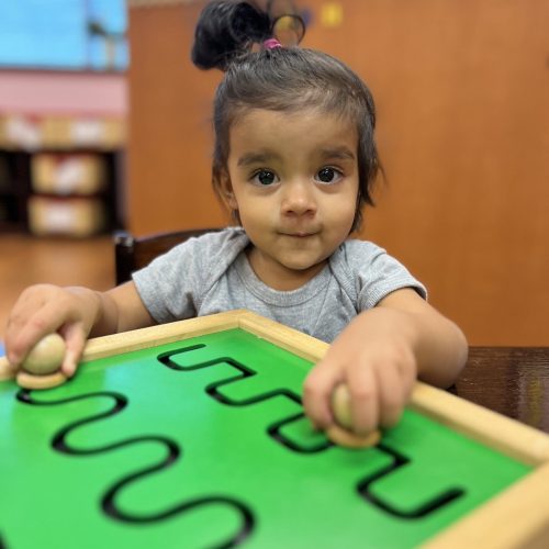 A toddler with dark hair in a bun is playing with a green wooden puzzle featuring wavy black lines. They hold wooden knobs and are sitting at a table in a classroom setting, with colorful shelves in the background.
