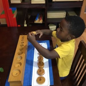 A young child wearing a yellow shirt is sitting at a table, engaging with a Montessori-style wooden cylinder block toy. The child is focused, placing the cylinders into matching holes. Wooden shelves with more educational materials are in the background.