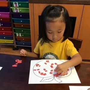 A child wearing a yellow shirt is sitting at a table, pasting red paper squares onto a drawing of an apple. In the background, there are colorful drawers and wooden cabinets.
