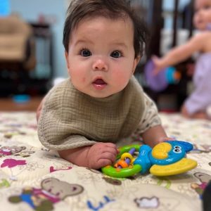 A baby with big eyes is lying on a patterned play mat, holding a colorful toy. The background is softly blurred, showing some furniture and a partially visible person. The baby looks curious and engaged.