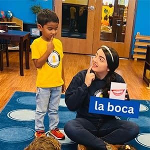A child and a teacher are sitting on a classroom floor. The child is wearing a yellow shirt, and the teacher is holding a flashcard labeled la boca with a picture of a mouth. Both are pointing to their mouths, engaging in a language activity.